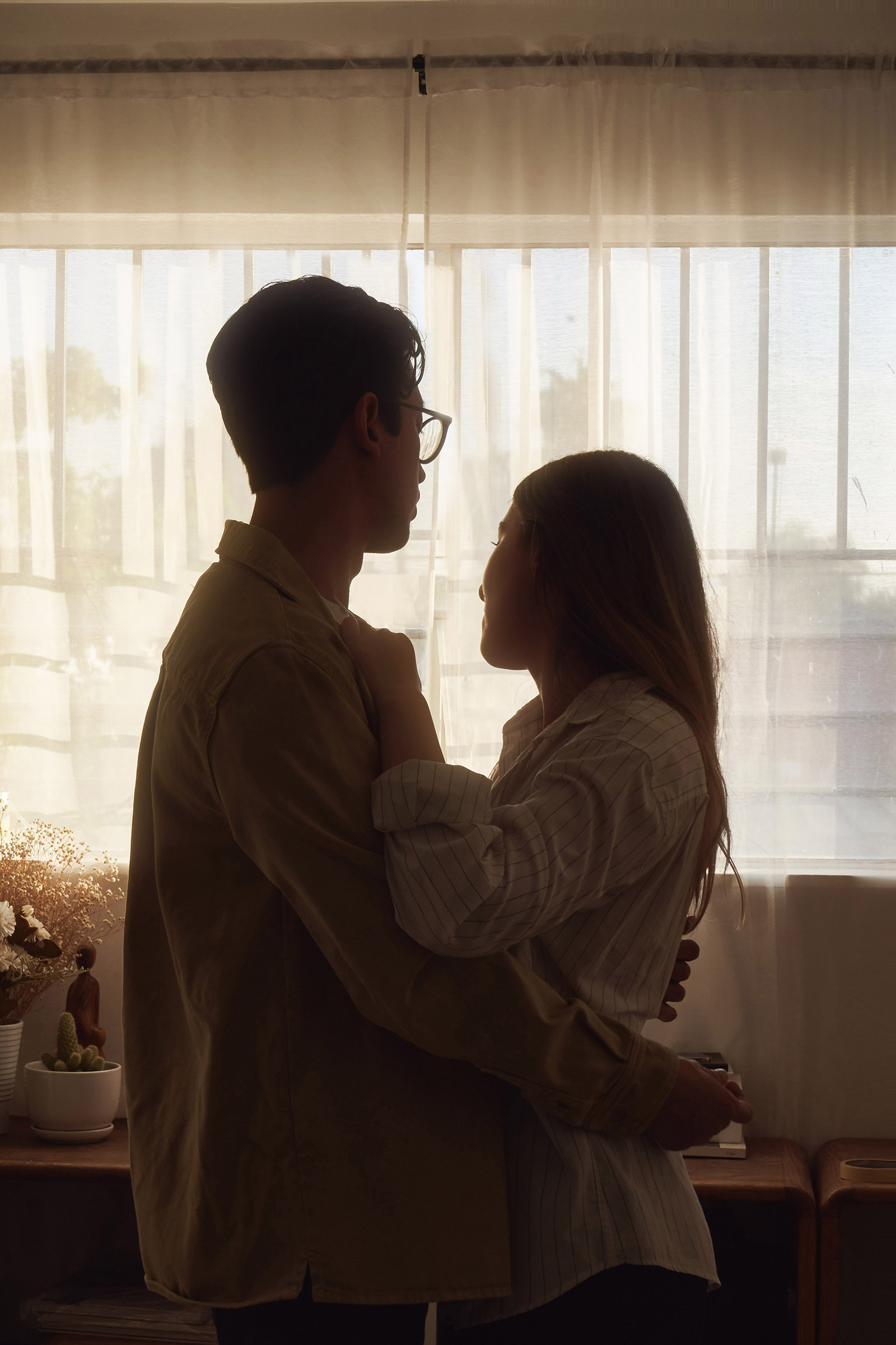 Gaby and Me a couple stand close by a window bathed in soft sunlight sharing an intimate moment.